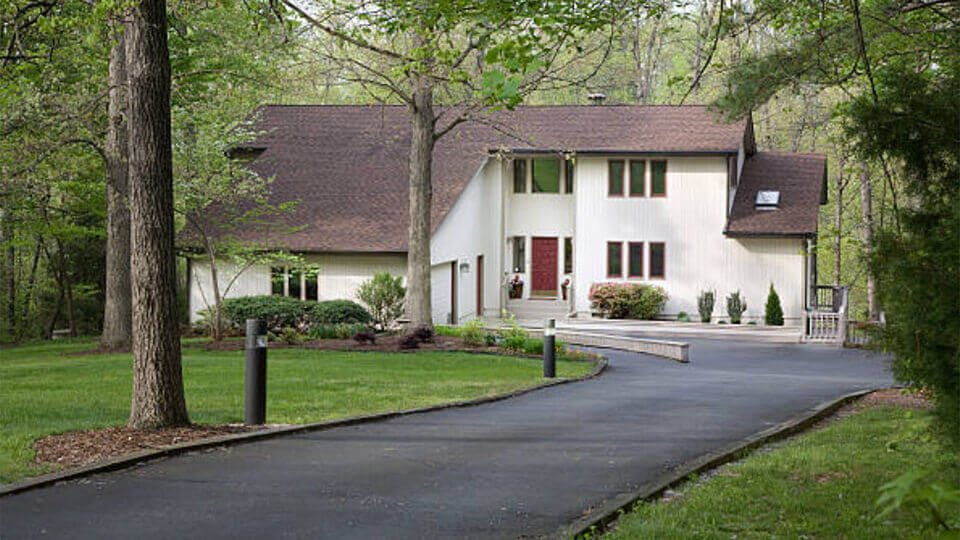 Modern two-story house with a sloped roof, surrounded by trees and a lawn, along a paved driveway.