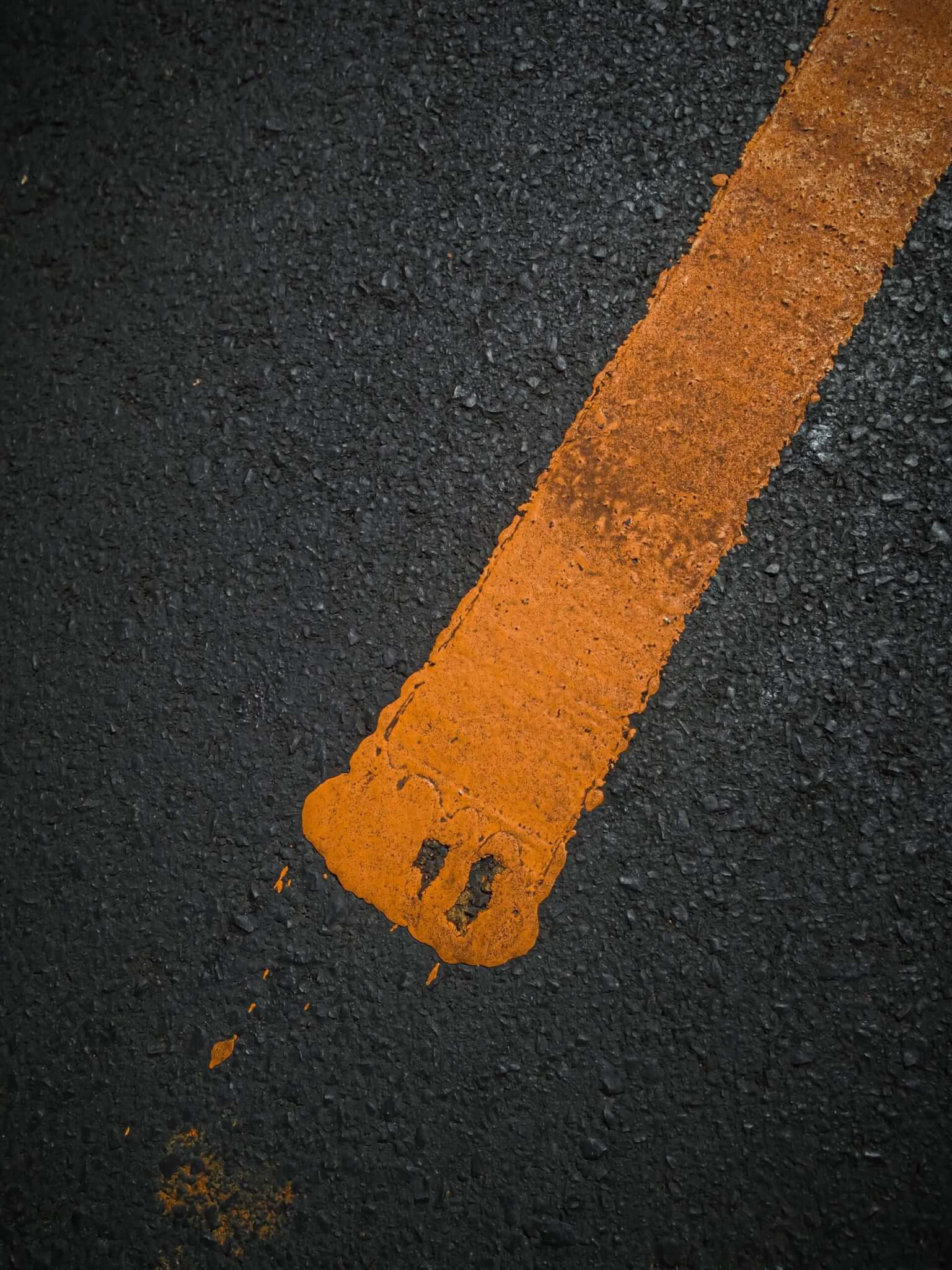Close-up of an orange paint line on dark asphalt, with some irregularities at one end.
