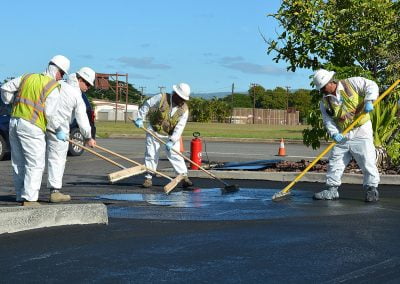 Under a clear sky, workers in safety gear diligently spread asphalt on the road with brooms, their expertise answering any asphalt sealcoating questions that arise.