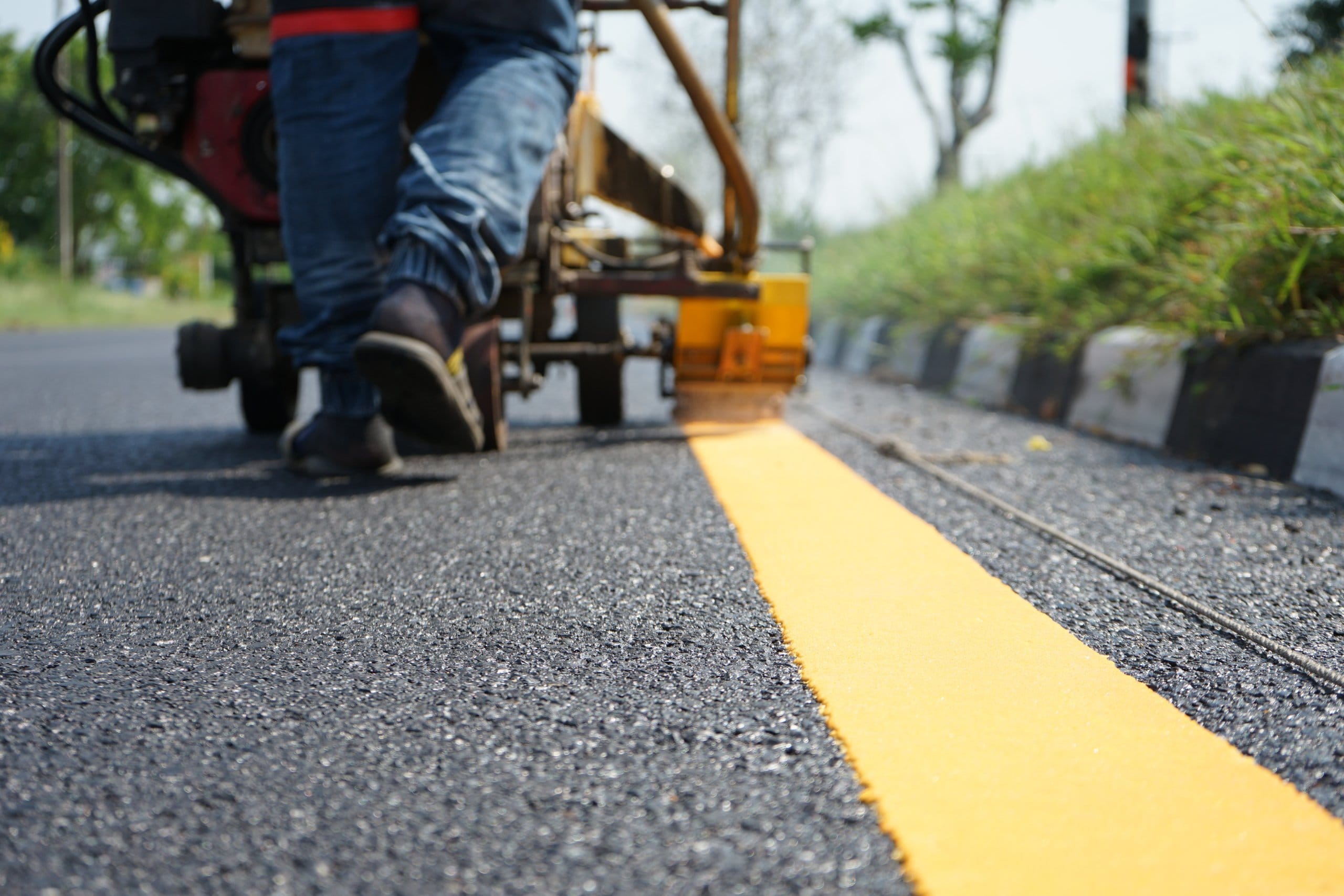 The officer was hitting a traffic line on a paved road. Worker uses a machine to paint a yellow line on a freshly paved road near a grassy curb.