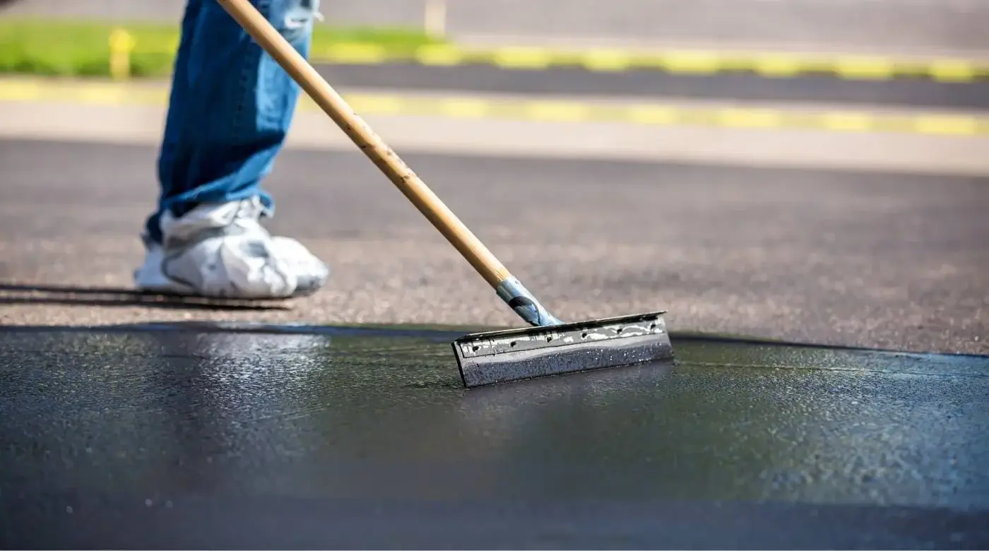 service-hero-image-9-1-1.jpg Person spreading sealcoat on asphalt with a squeegee, wearing jeans and white sneakers.