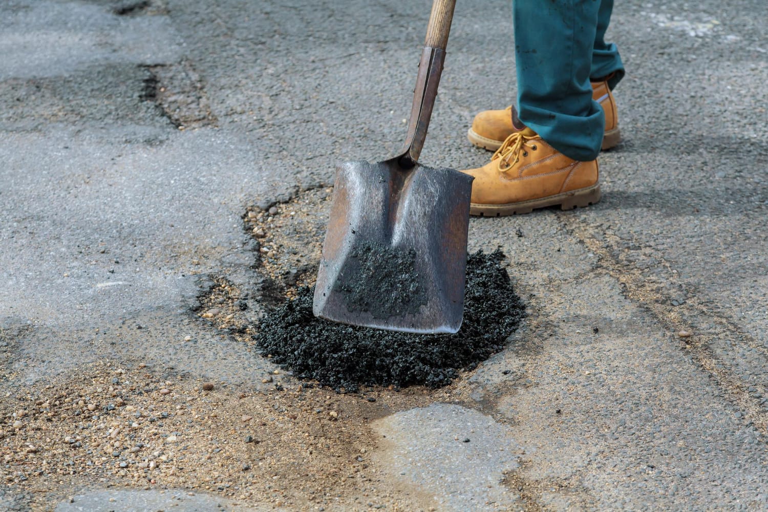 3468831 A person in work boots fills a pothole in asphalt using a shovel.