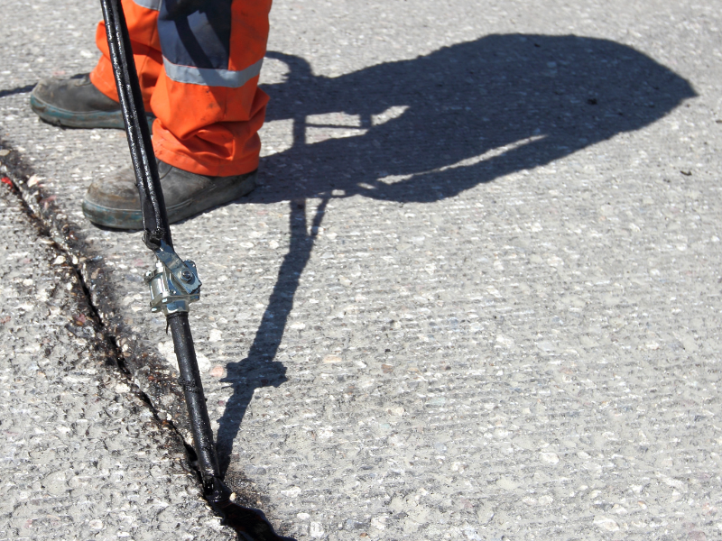 Worker in orange pants sealing a crack in pavement with a tool on a sunny day.