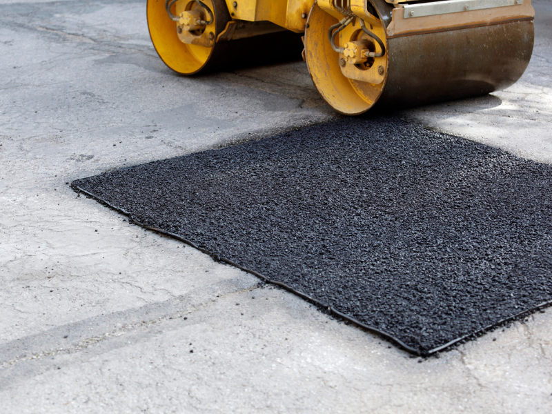 Yellow steamroller compacting fresh asphalt patch on a concrete surface.
