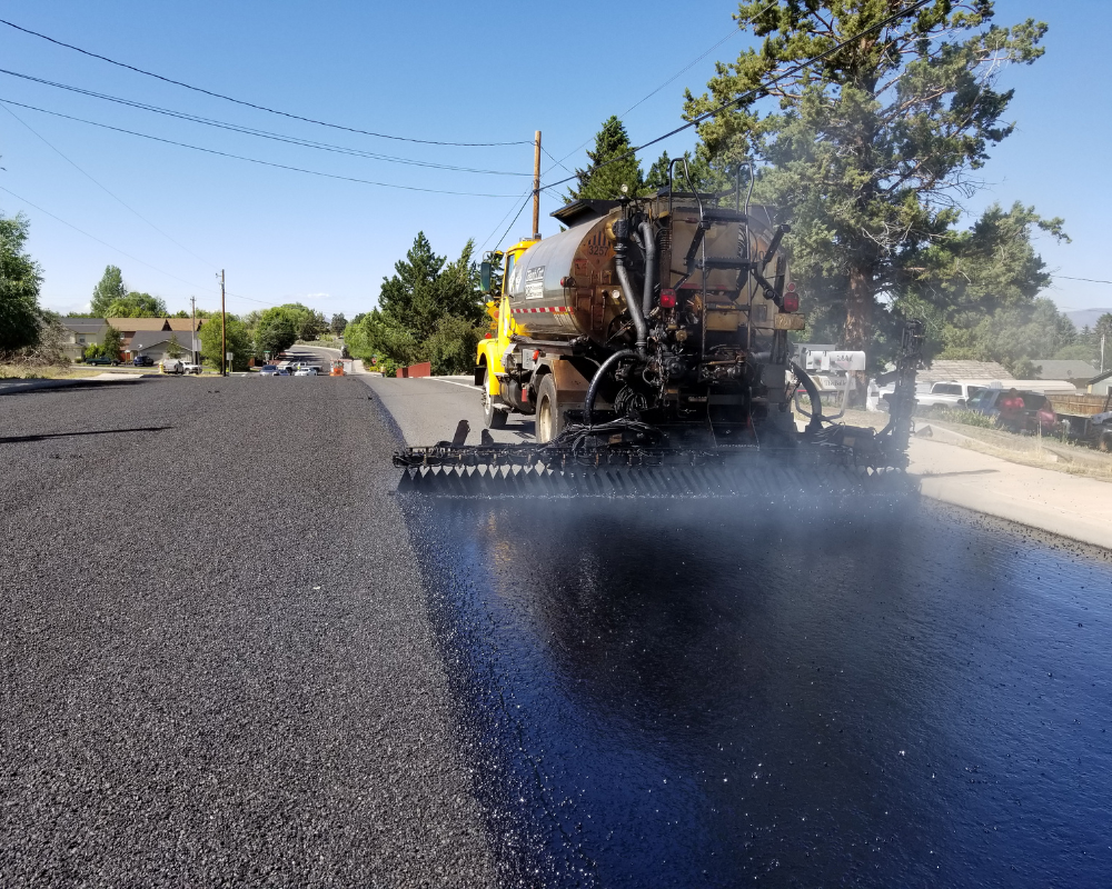 Asphalt Sealcoating A truck sprays black asphalt on a road during paving, with steam rising and trees in the background.