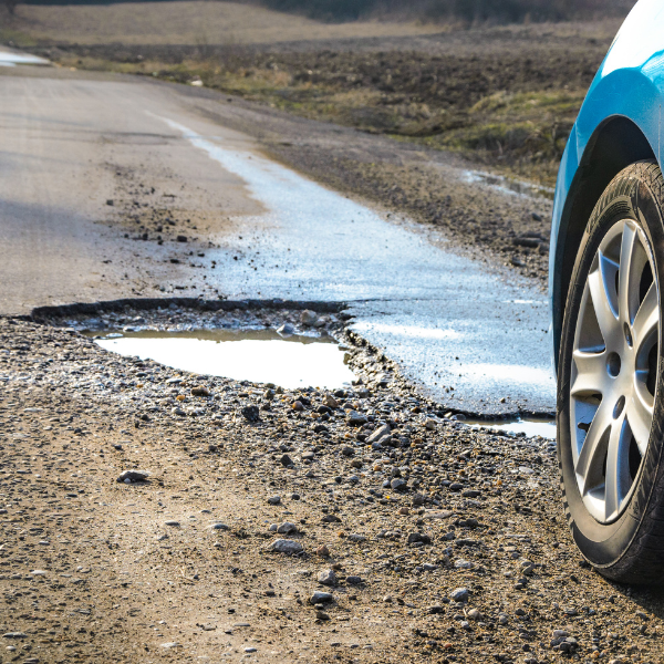 PotHole Repair (1) A blue car near a large pothole filled with water on a rural asphalt road.