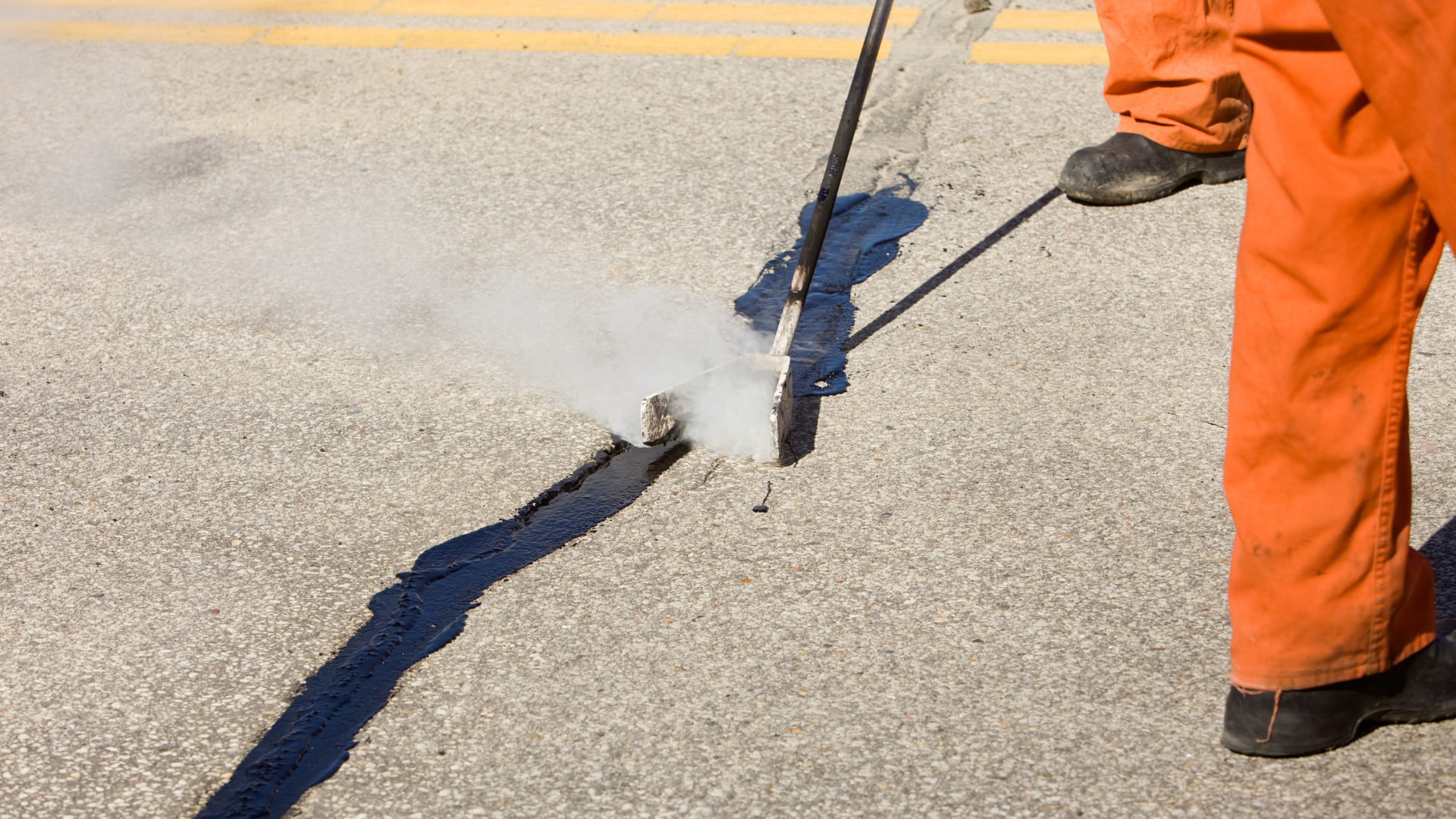 Worker in orange pants sealing a road crack with hot material, steam rising from the crack.