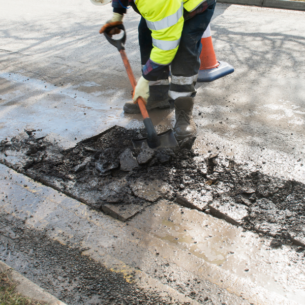 pothole Repair (2) Worker in a high-visibility jacket uses a shovel to dig up broken asphalt on a road.