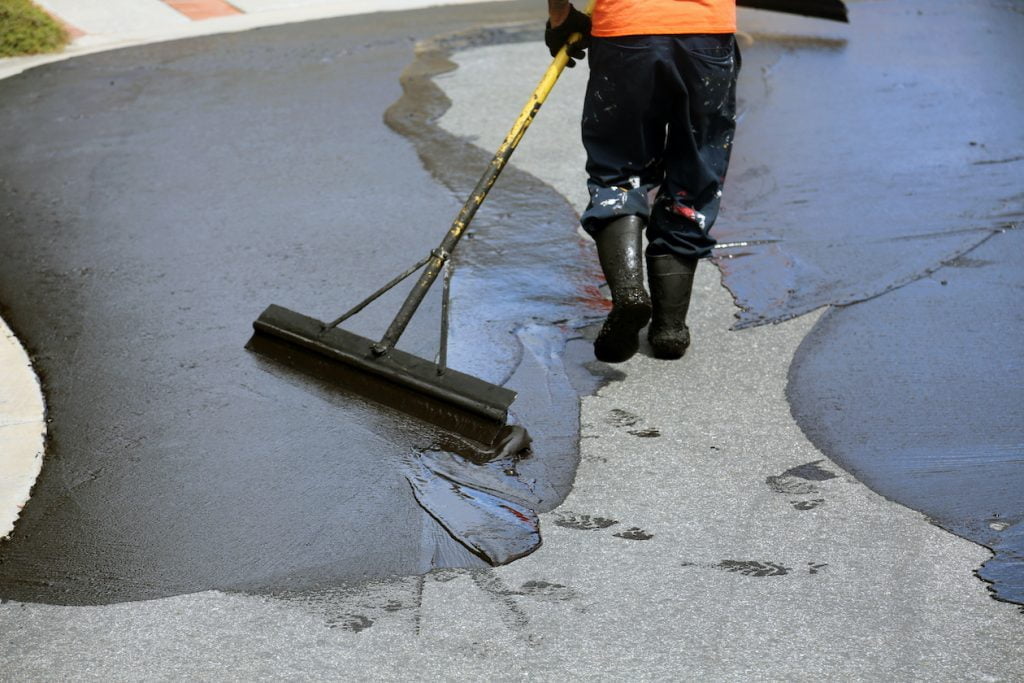 A worker spreads black sealant on a driveway with a large squeegee, wearing boots and protective clothing.