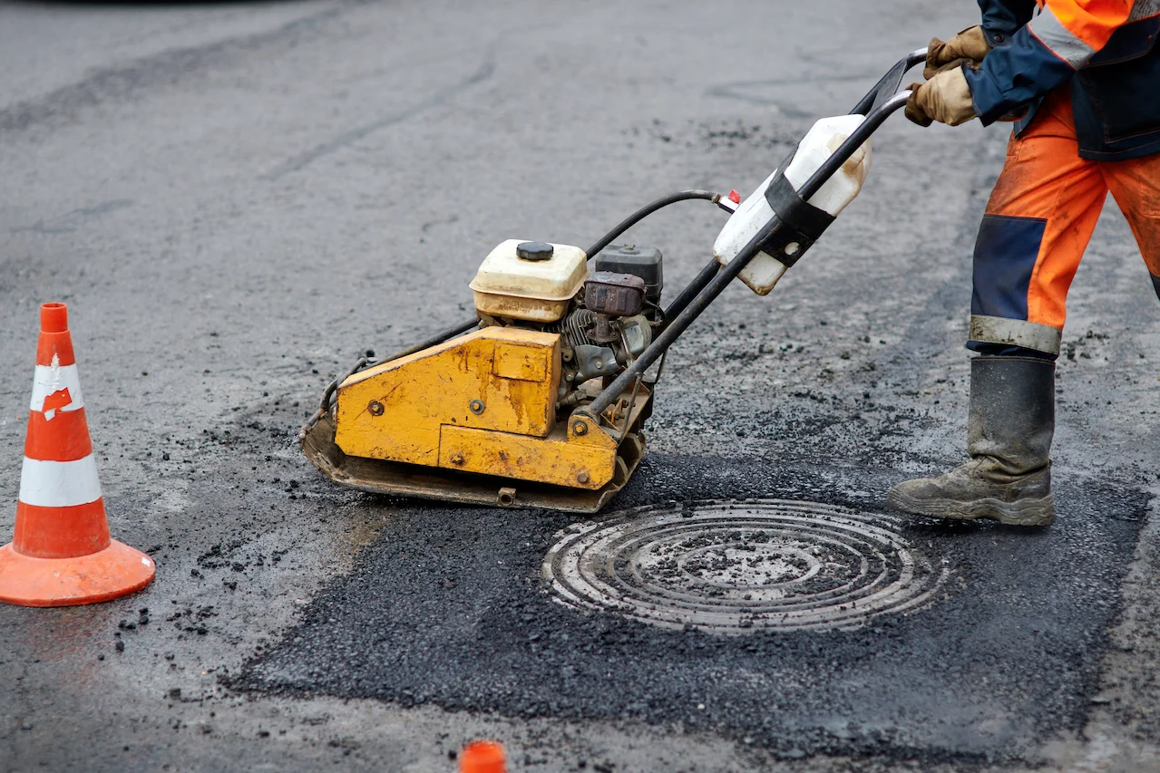 Asphalt-PavementServices.jpg A worker uses a yellow plate compactor to repair asphalt near an orange traffic cone on a road.